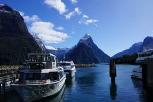 A photo of Milford Sound. There are small cruising boats on the left of the picture with snow capped mountains in the background. The sky is blue with just a few white clouds. The sunshine reflects on the surface of the lake.