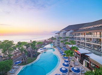 Resort with balconies overlooking curved pool and beach at sunset with blue umbrellas and lounge chairs Caption: The image shows a large resort with multiple floors and balconies overlooking a curved swimming pool. There are numerous blue umbrellas and lounge chairs arranged around the pool area. The resort is situated next to a beachfront, with the ocean visible in the background during sunset. The sky is clear with soft colors from the setting sun.