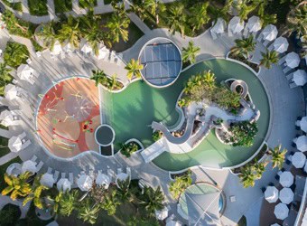 Aerial view of a pool complex with water playground, slide, sun loungers, umbrellas, and palm trees.