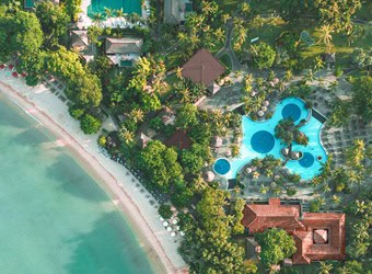 erial view of a coastal resort with a sandy beach, pool, palm trees, and multiple buildings.