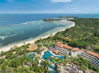 Aerial view of a coastal resort with red-tiled roofs, large pool, beach, and peninsula.