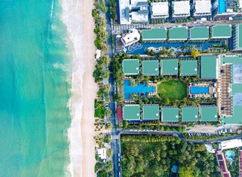 Aerial view of beach with turquoise ocean, adjacent road, and beachfront buildings with pools and greenery.