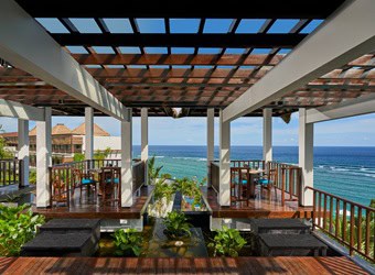 Wooden deck with shaded seating, water feature, and ocean view with coastal background. Caption: The image shows a wooden deck with shaded seating areas overlooking the ocean. The structure features wooden beams and railings, with a small water feature and greenery in the center. The view extends to the clear blue sea and a coastal area in the background.