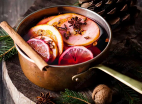 A copper pot of mulled wine with orange and apple slices, cinnamon sticks, and star anise, sits on a wooden surface, evoking a cozy, festive atmosphere.